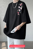 Person wearing a black oversized shirt with floral embroidery, holding a phone, against a gray background.