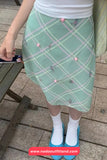Person wearing a green plaid skirt with pink buttons on a sidewalk.