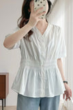 A woman takes a mirror selfie wearing a white short-sleeve button-up blouse with a smocked waist and peplum hem, paired with light blue pants.
