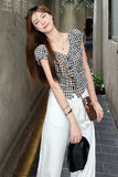 Woman in a checkered top and white pants standing on a sidewalk.