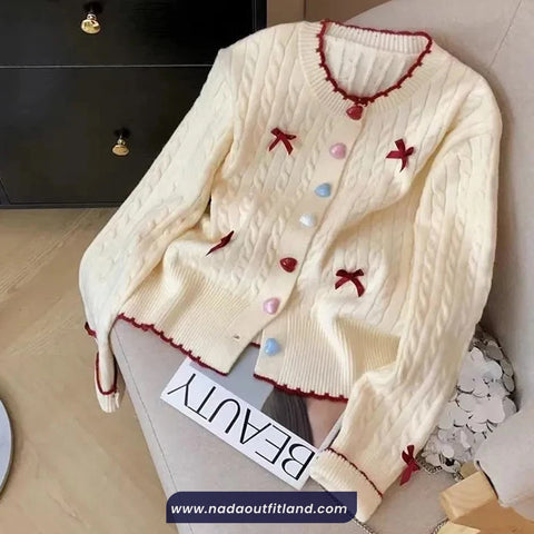 White cardigan with decorative bows and buttons on a chair, branded 'Nada Outfit Land'.