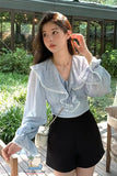 Woman wearing a light blue blouse with ruffled details and black shorts outdoors.