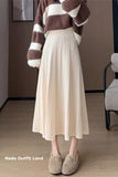 Person wearing a beige pleated skirt and brown sweater in a room.