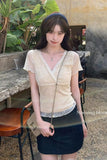 A woman wearing a beige short-sleeve lace-trim V-neck top and a dark denim mini skirt, standing outdoors in a cafe-like setting with plants and rustic furniture.