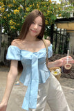 Woman in a light blue off-shoulder top and white pants holding a drink, standing outdoors with greenery.
