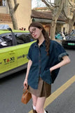 A woman stands on a city street wearing a dark blue denim short-sleeve button-up shirt, a brown mini skirt, and glasses. A bright yellow taxi is visible behind her.