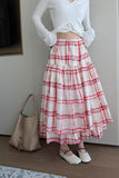 Person wearing a white long-sleeve top and red plaid skirt with a woven bag on a neutral background