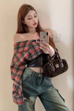 Woman holding a phone and a handbag, wearing an off-shoulder plaid top and jeans.