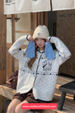 Woman wearing a white sweater with black patterns, beige knit hat, and blue gloves, sitting on a wooden bench.
