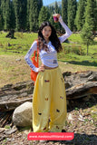 Person in a white top and yellow pants standing outdoors with trees and rocks in the background.