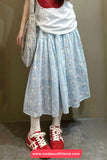 Person wearing a light blue floral skirt with a white top and red shoes, standing against a plain background.
