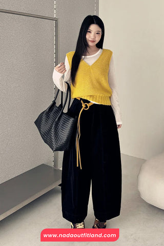 Woman wearing a yellow vest and black pants holding a black bag indoors.