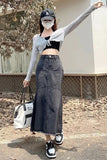 A woman posing outdoors wearing a gray long-sleeve crop top, a black lace-up sweetheart bustier underneath, a dark gray denim maxi skirt, a black baseball cap, a white face mask, and carrying a white tote bag.