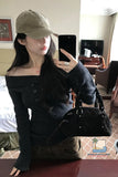 Person wearing a black off-shoulder sweater and beige cap, holding a black handbag in a room.