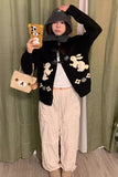 Person wearing a black cardigan with bunny patterns, holding a phone with a bear case, standing against a green curtain.