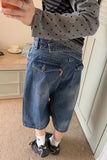 A person wearing blue denim cropped shorts with a matching denim belt, paired with a gray polka-dot long-sleeve top, white socks, and black sneakers in an indoor room setting.