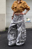 Person wearing a yellow plaid shirt and camouflage pants standing in front of a white wall.
