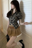 Woman wearing a plaid shirt and beige skirt standing against a wooden panel wall.