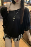 A person wearing a loose black off-the-shoulder t-shirt and black-and-white striped shorts, accessorized with a silver cross necklace in an indoor setting.