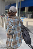 Person wearing a plaid coat and cap, holding a black bag, in an outdoor setting.