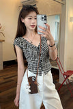 Woman taking a mirror selfie wearing a black and white checkered top and white pants.