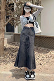 A woman stands outdoors wearing a gray knit long-sleeve crop top with a black bow detail, a black crop top underneath, a long dark gray denim skirt with front patch pockets, a white tote bag, and black-and-white sneakers.