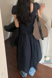 Person wearing a dark checkered dress with a beige bag, standing indoors.