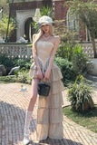 Woman in a beige dress and cap standing outdoors with greenery and a building in the background