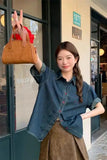 A woman stands outdoors in front of a storefront, holding up a tan leather handbag. She is wearing a blue denim button-up shirt and a brown skirt.