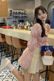 Woman in a pink top and white skirt holding a phone and white handbag in a cafe.