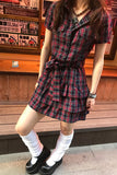 Person wearing a red and blue plaid dress with white socks and black shoes in front of a wooden wall with posters.