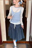 Person wearing a light blue sweater over a white shirt with a plaid skirt, standing in a doorway.