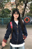 A young woman wearing a dark blue denim jacket with red plaid accents on the cuffs and collar, layered over a white t-shirt and light blue jeans. She is standing outdoors in front of trees and a railing.