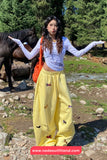 Person wearing a white long-sleeve top and yellow pants with animal prints, standing outdoors with horses in the background.