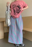 Person wearing a pink t-shirt with a graphic design and a long plaid skirt, holding a silver bag indoors.