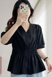 A woman takes a mirror selfie showing a black short-sleeve blouse with a V-neck, button-up front, and a gathered peplum waist. The blouse has a subtle sheen and is paired with light blue jeans.