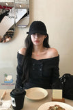 Woman sitting at a table with a black coffee cup and plates, wearing a black off-shoulder top and black cap.
