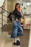 A woman takes a mirror selfie in an indoor setting, wearing a black and gray striped long-sleeve top over a sheer gray polka-dot long-sleeve layer, paired with wide-leg blue denim shorts, black sneakers, and a black backpack.