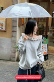 Woman holding a transparent umbrella in an urban setting, wearing a light gray sweater with ruffled details.