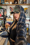 Person wearing a plaid jacket and cap in a casual setting with shelves and a refrigerator in the background.