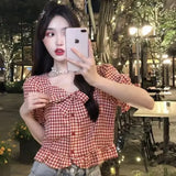 Woman taking a selfie in an outdoor setting with a checkered top and pearl necklace.