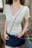 A woman wearing a cream-colored lace short-sleeve top with a V-neck and ruffled sleeves, paired with a dark denim mini skirt. She is carrying a silver chain strap bag and wearing delicate jewelry, standing in a rustic indoor setting with wooden floors and walls.