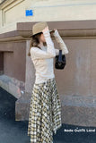 Woman adjusting her hat against a building wall, wearing a beige top and plaid skirt.