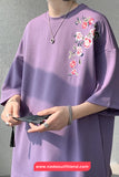 Person wearing a purple oversized sweatshirt with floral embroidery, holding a phone against a gray background.