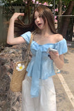 Woman in a light blue top and white pants holding a phone and a woven bag with a flower, standing outdoors.