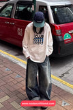 Person wearing a beige sweatshirt with graphic design and blue jeans standing on a sidewalk with a taxi in the background.