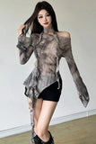 A woman modeling a sheer, asymmetrical long-sleeve top with a gray and brown mottled pattern, paired with black shorts and black boots, standing against a plain indoor wall.
