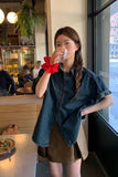 A young woman stands in a cafe wearing a dark blue denim shirt with rolled-up sleeves and a brown skirt, holding a clear plastic cup to her mouth. She has a bright red scrunchie on her wrist.