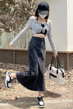 A woman wearing a gray long-sleeve crop top and a dark denim maxi skirt, carrying a white tote bag and wearing a black cap and face mask. She is walking outdoors on a paved path with dry leaves and a fence in the background.