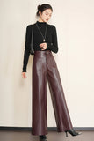 A woman modeling a black long-sleeve mock-neck top and high-waisted, wide-leg burgundy faux-leather pants, standing against a light beige wall.